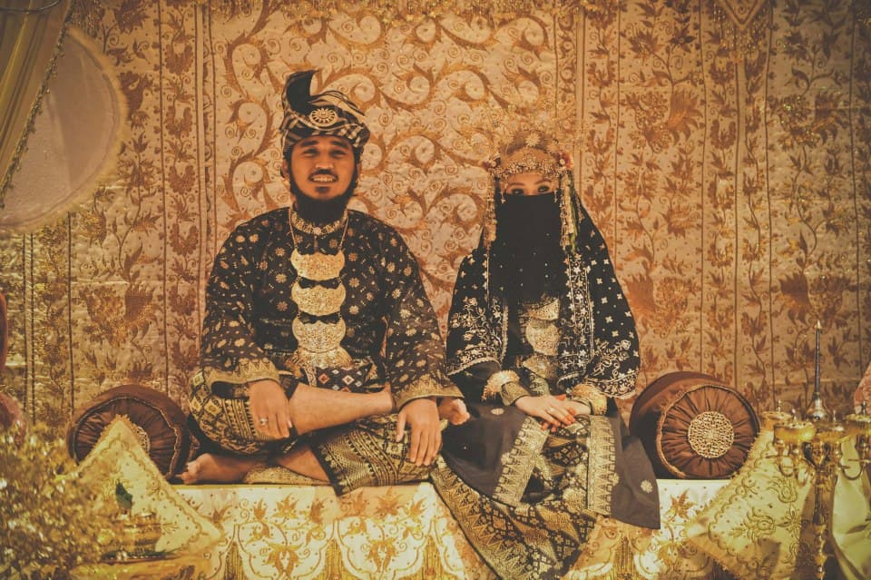 A Malay bride and groom in gold-embroidered ceremonial songket seated side by side on the pelamin during the bersanding