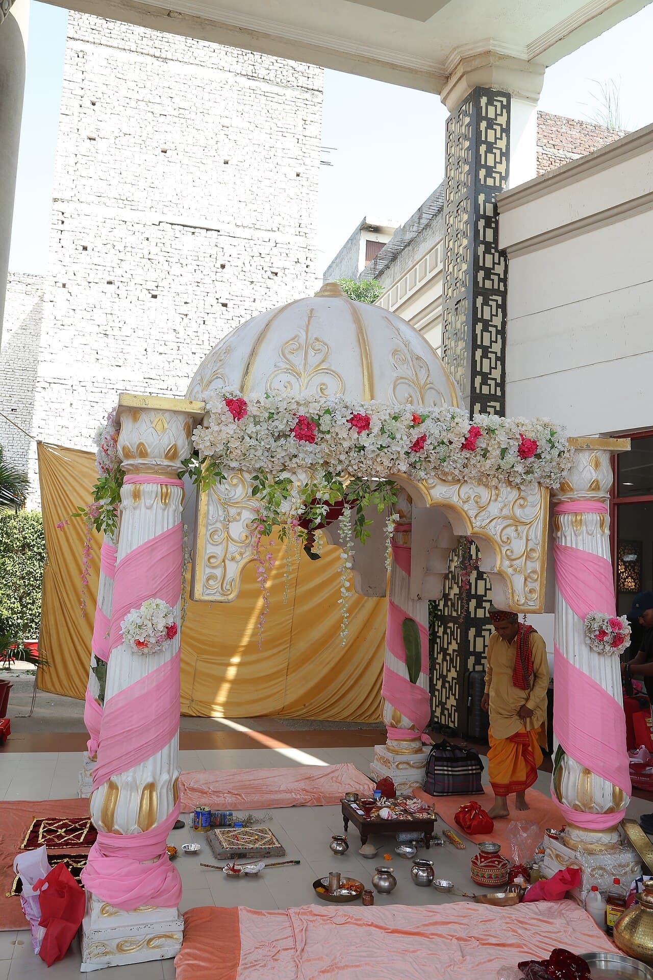 A decorated Hindu wedding mandap — flower-draped pink-and-white pillars under a gold canopy, with ritual trays laid out in front