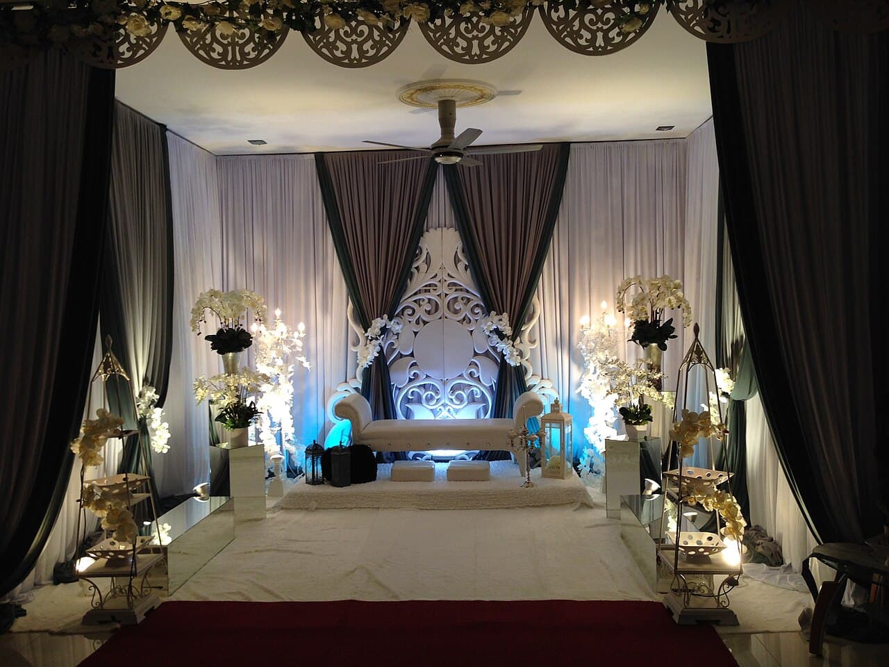 A white-and-grey Malay pelamin (decorated wedding dais) with a carved throne seat, orchids and draped curtains